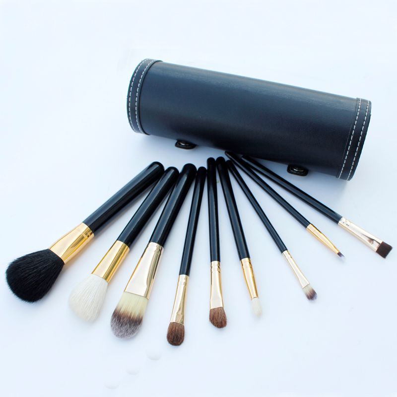 Set of makeup brushes with black handles and gold accents, arranged in front of a black cylindrical case on a light blue background.