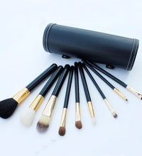 Set of makeup brushes with black handles and gold accents, arranged in front of a black cylindrical case on a light blue background.